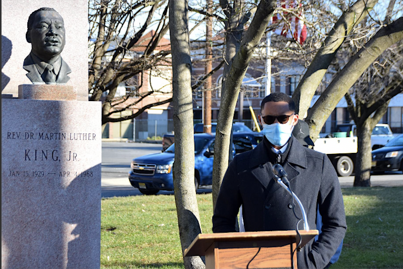 Tymek Jones with mask and sunglasses speaking at podium next to statue of Martin Luther King Jr. outdoors