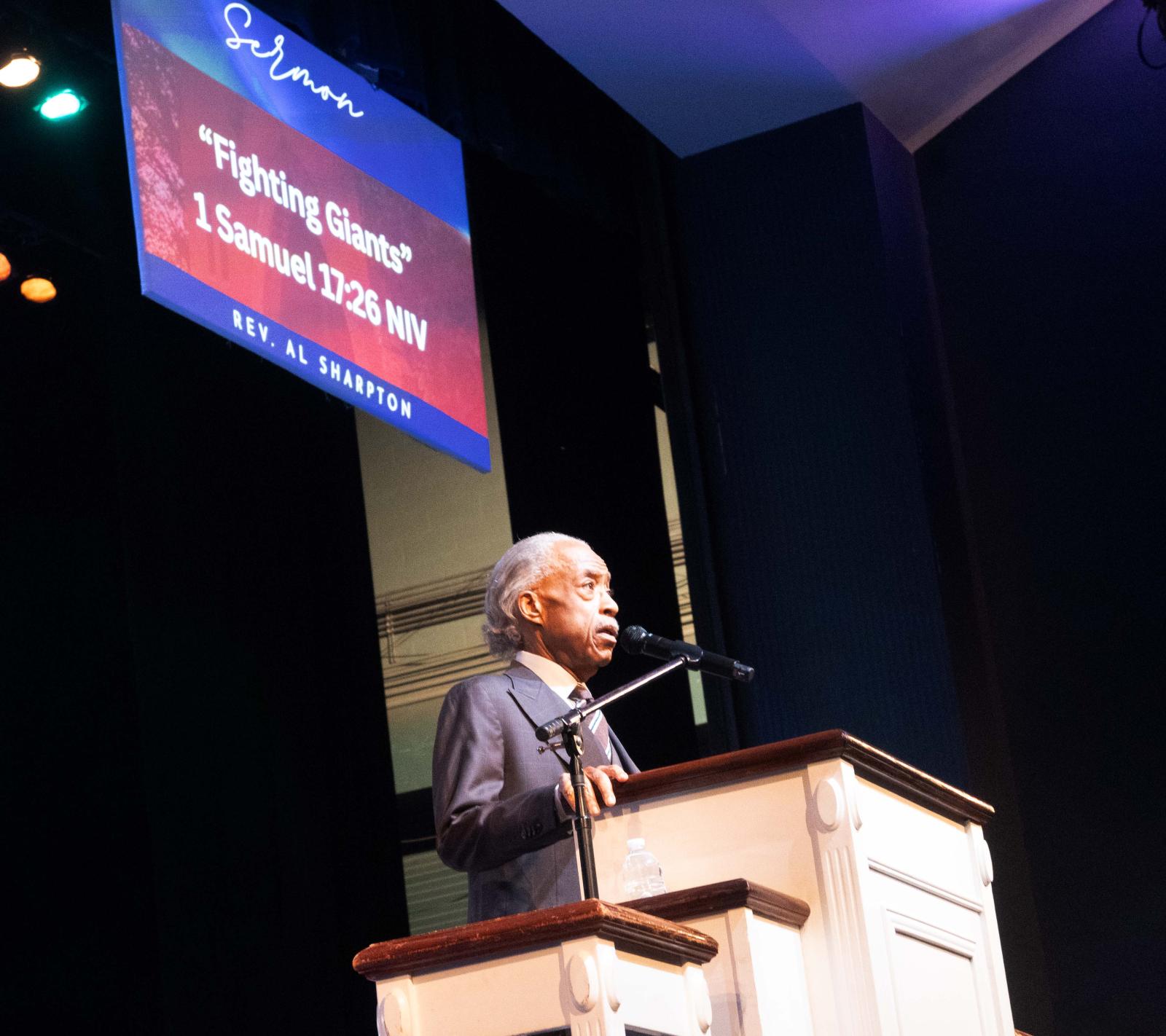 Man standing behind a pulpit delivering remarks. There is a screen at the top left of the image that says "Fighting Giants" 1 Samuel 17:26 NIV