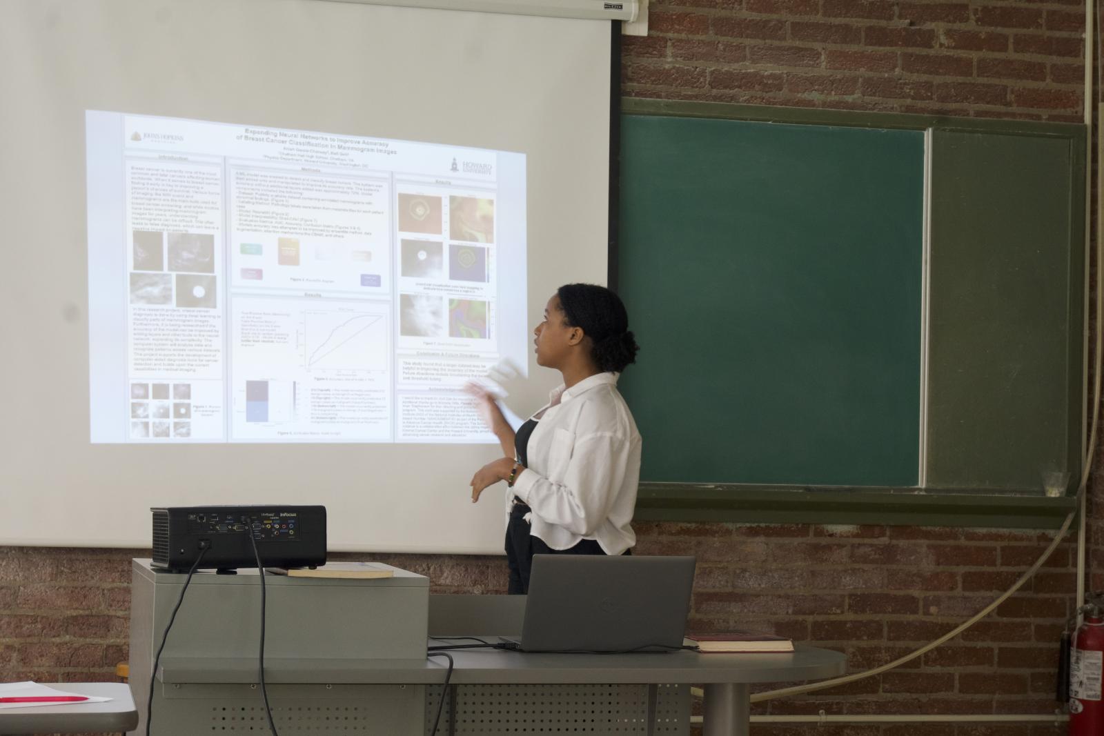 High school student looking at research presentation that is projected onto a screen. She is in a classroom. there is a green chalkboard in the background.