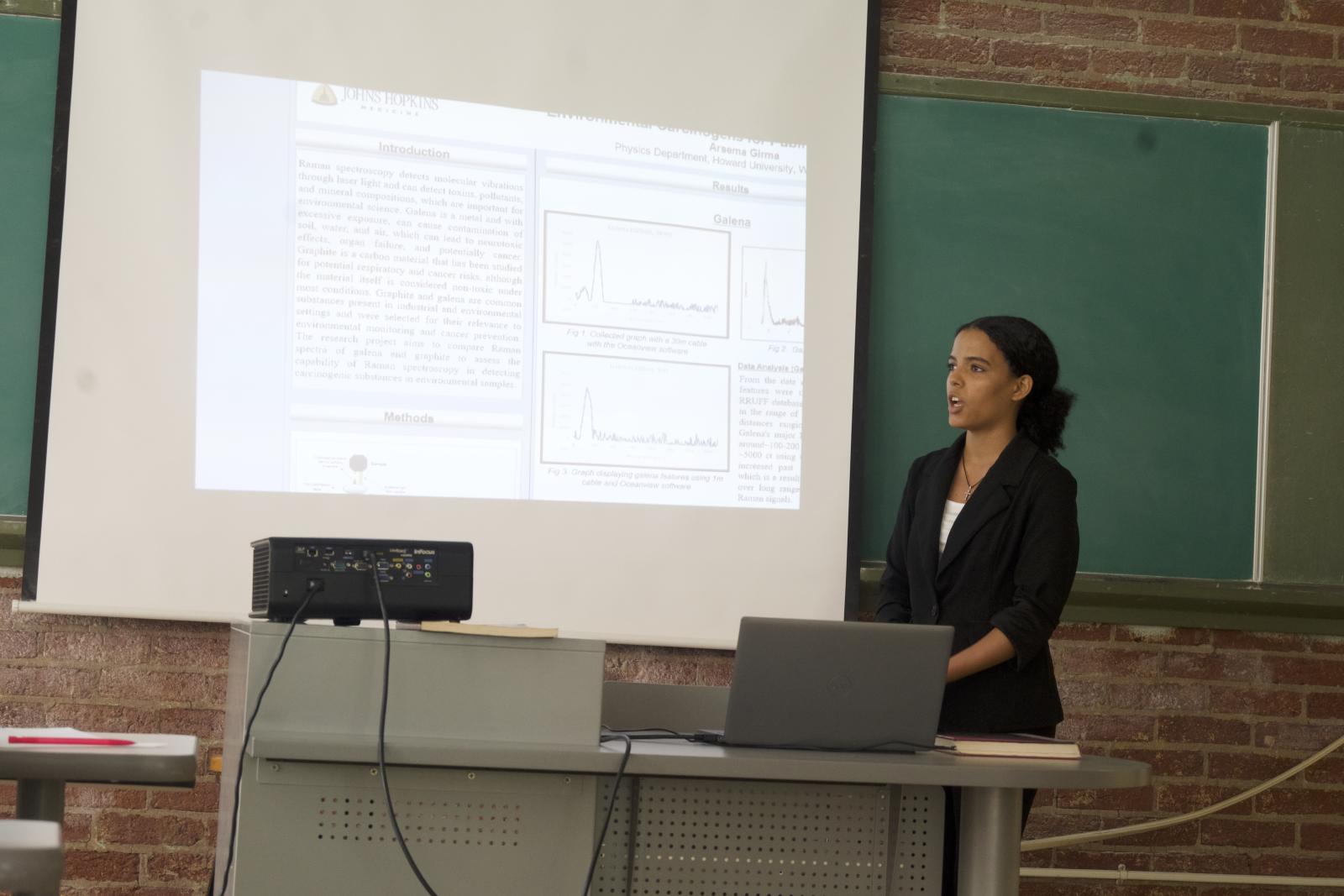 High school student in mid sentence explaining her research findings. The research is projected onto a screen that is in front of a green chalkboard.