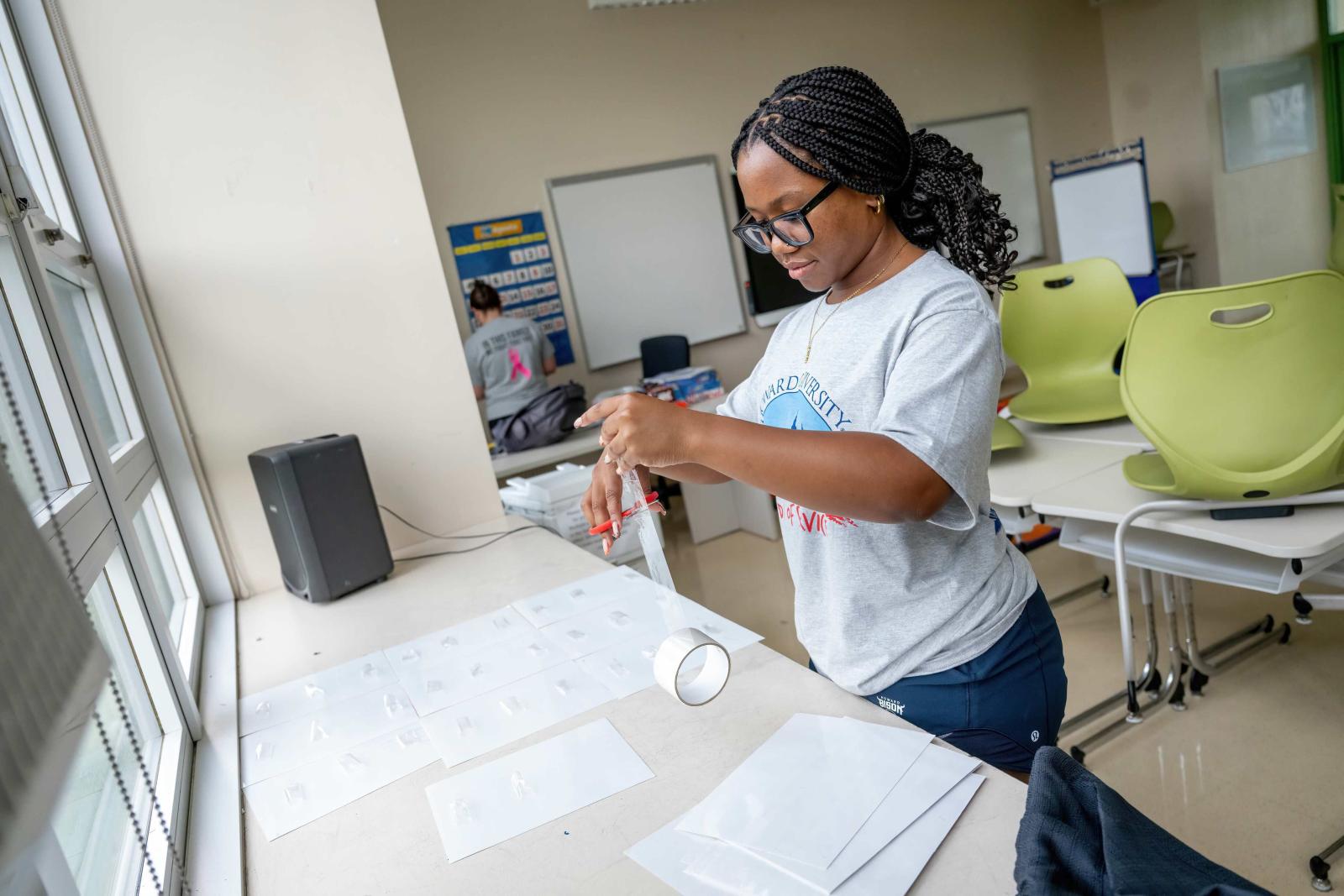 A college student is standing in a classroom of an elementary school. She is cutting strips of clear tape. There are pieces of paper on the desk in front of her with strips of tape on them as well