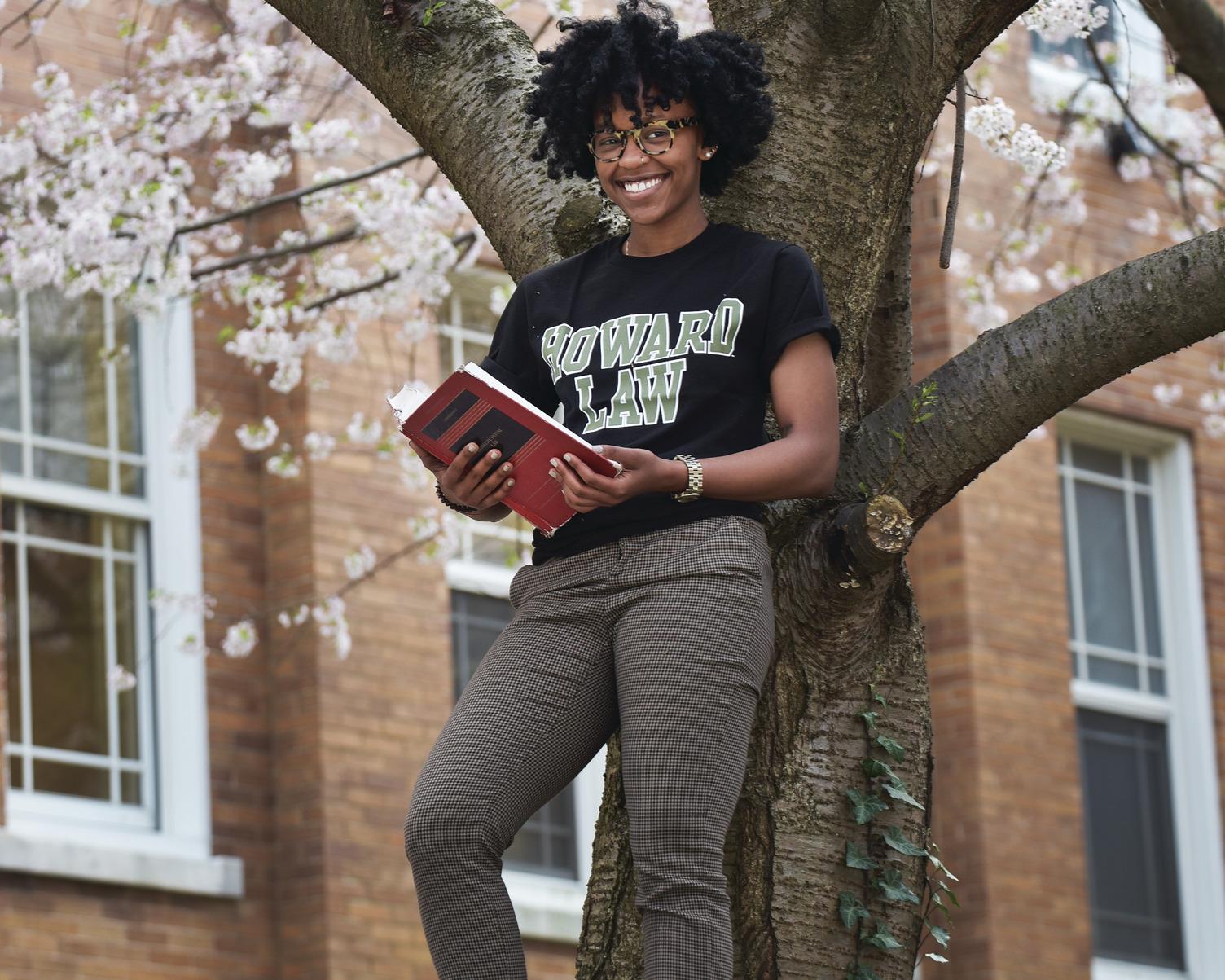 A Howard Law School student standing against a tree while holding a book