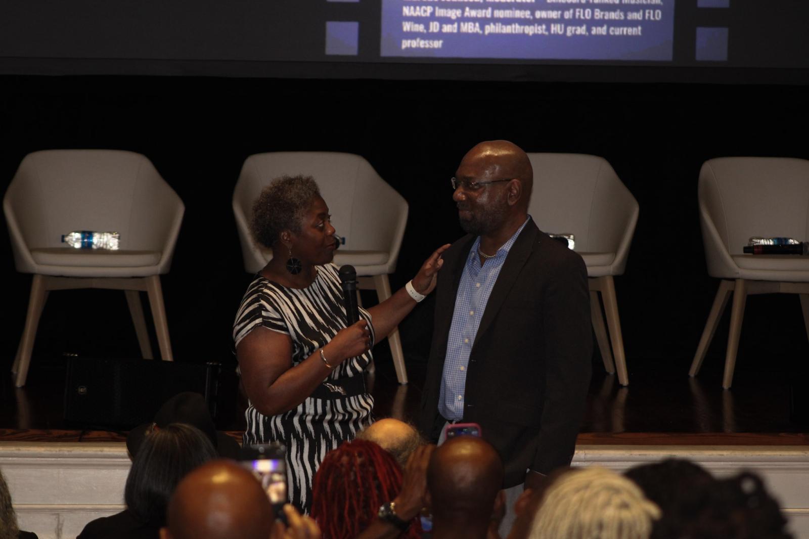 Two people standing in front of a stage that has four white chairs placed on the stage. The woman on the left is holding a microphone and has her hand placed on the man on the right's shoulder
