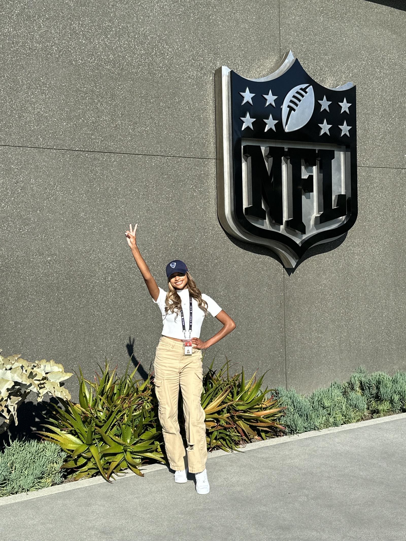 Woman standing in front of the NFL headquarters. There is a large NFL logo sign on the building in the back. The woman is holding up a peace sign