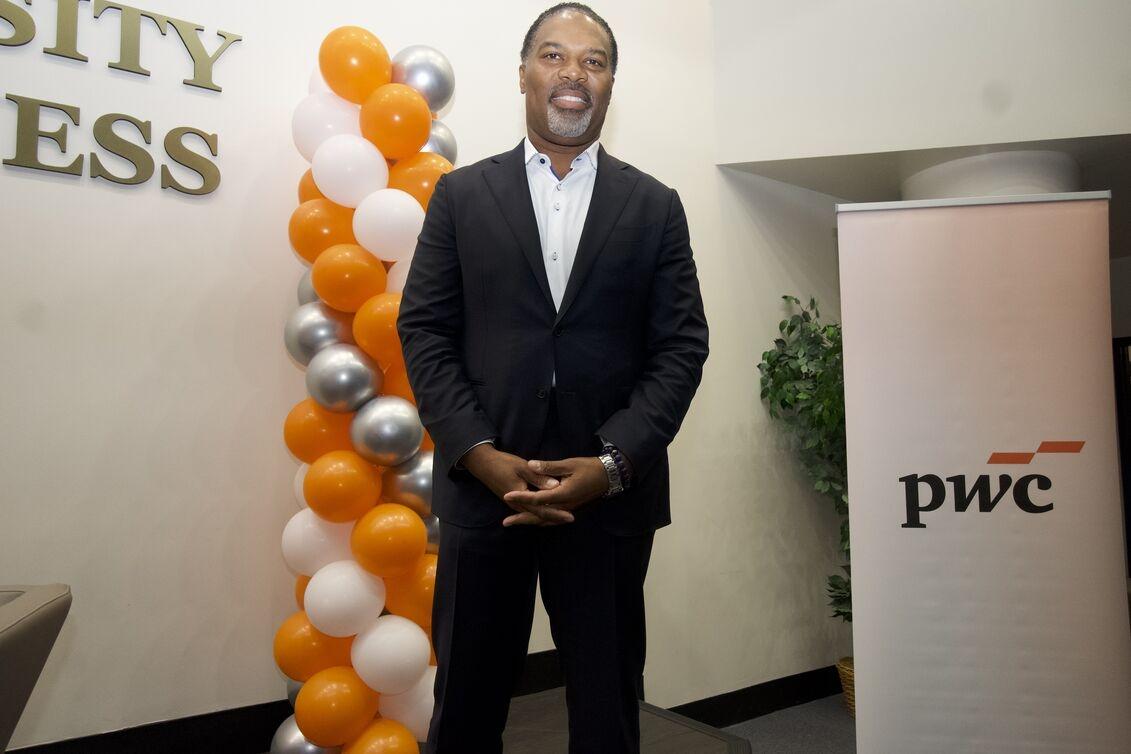 A man smiling at the camera in front of a structure made of different colored ballons. On the right of the man is a banner that says PwC
