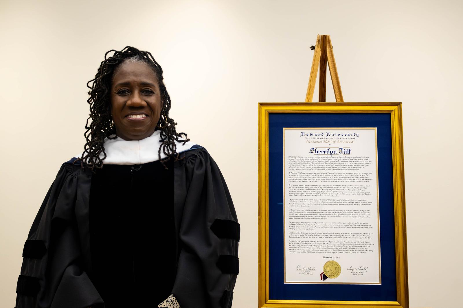 Woman standing in black university regalia (a black graduation-styled robe) with a white collar. She is standing next to an award from Howard University, the Presidential Medal of Achievement