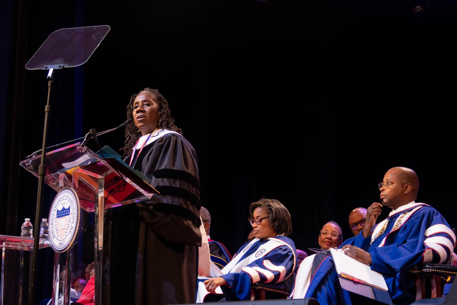 Several people on stage during a ceremony. There is a woman that is standing behind a podium with higher education regalia on. There are people seated behind her in higher education regalia on as well
