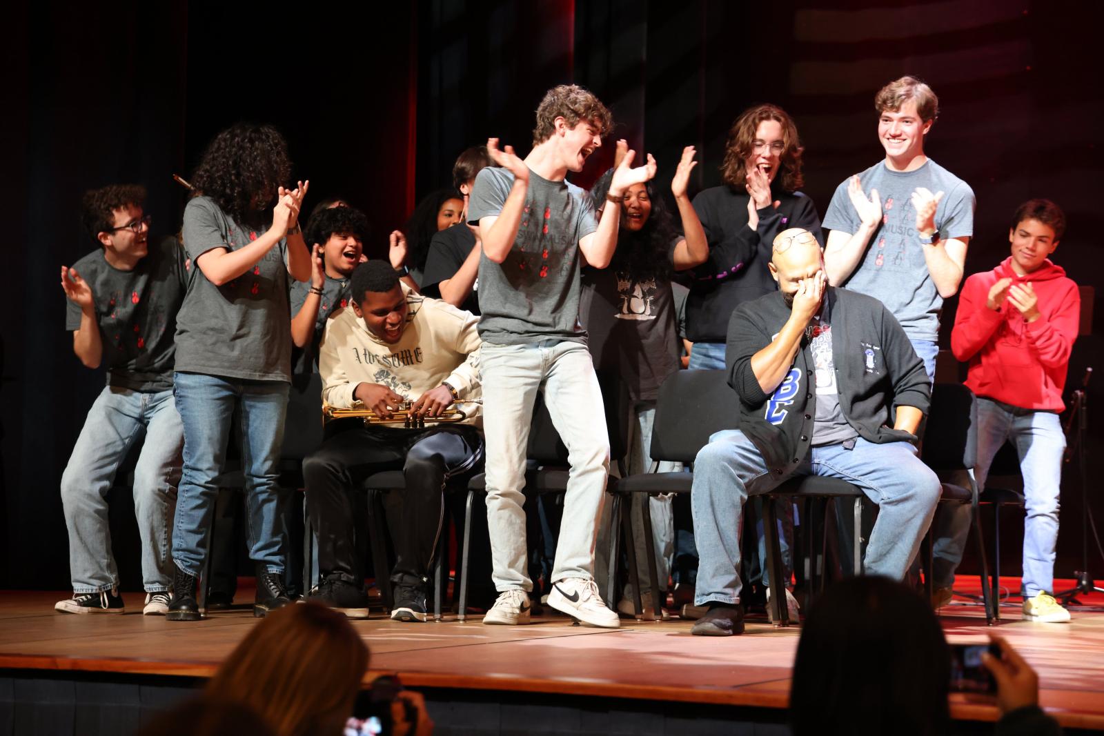 a group of people on stage, one is sitting in a chair, the others are all on their feet cheering and applauding