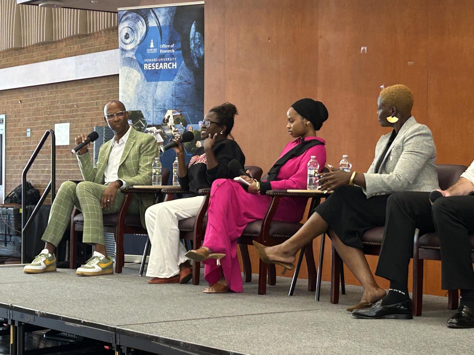 Four people sitting in chairs on stage. The two people on the left are holding microphones close to their mouths