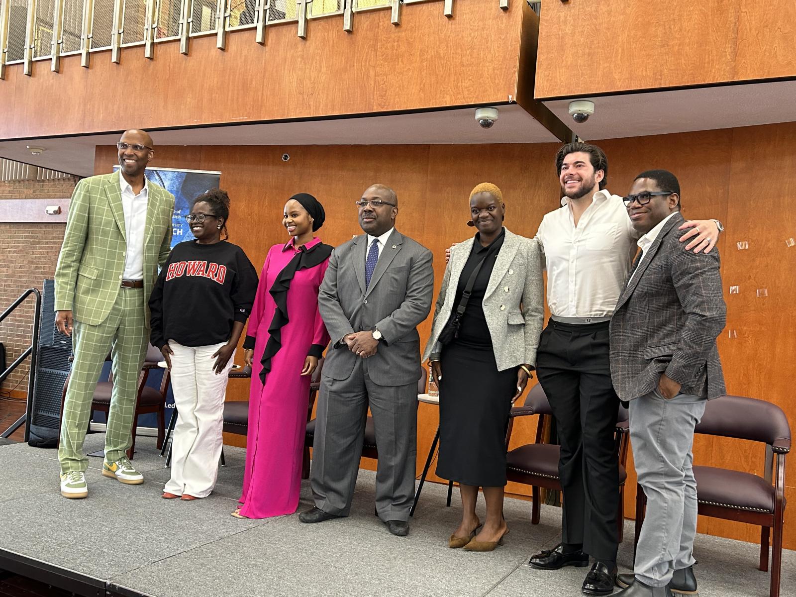 A group of seven people standing on a stage posing for a picture. The second to last person on the right has his hands over the shoulders of the person on the right and left of him