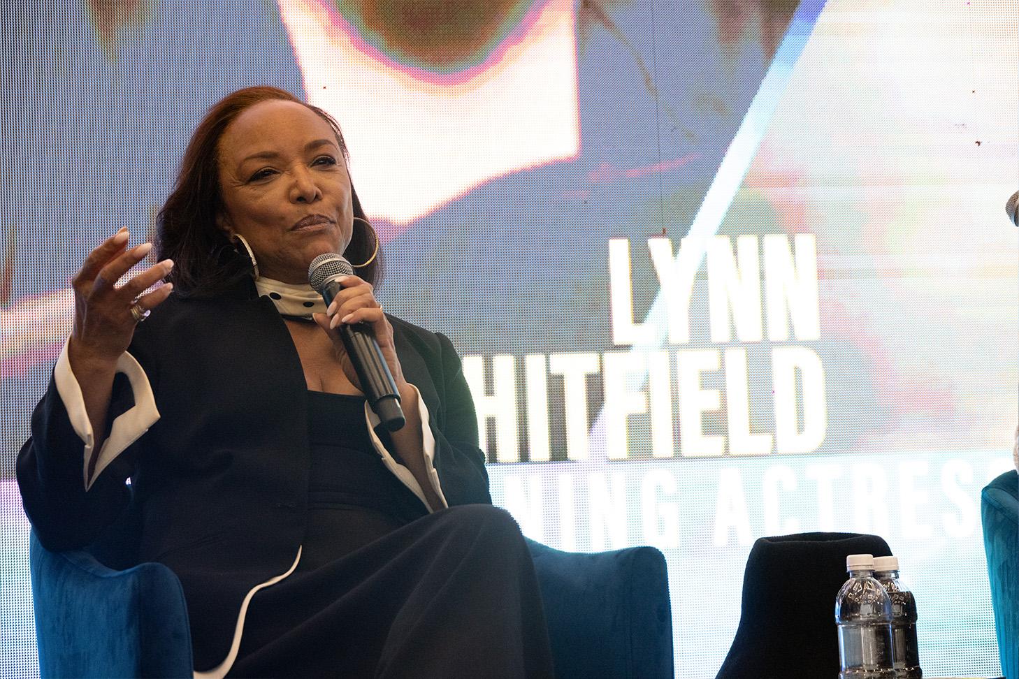 Actress Lynn Whitfield sitting on stage. She has a microphone in her right hand and is gesturing with her left hand