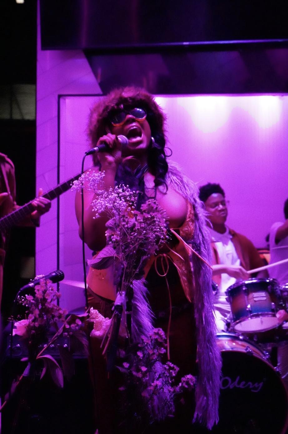 A performer who is holding a mic attached to a stand and has her mouth is open as she is in mind singing. there is a person playing drums behind her