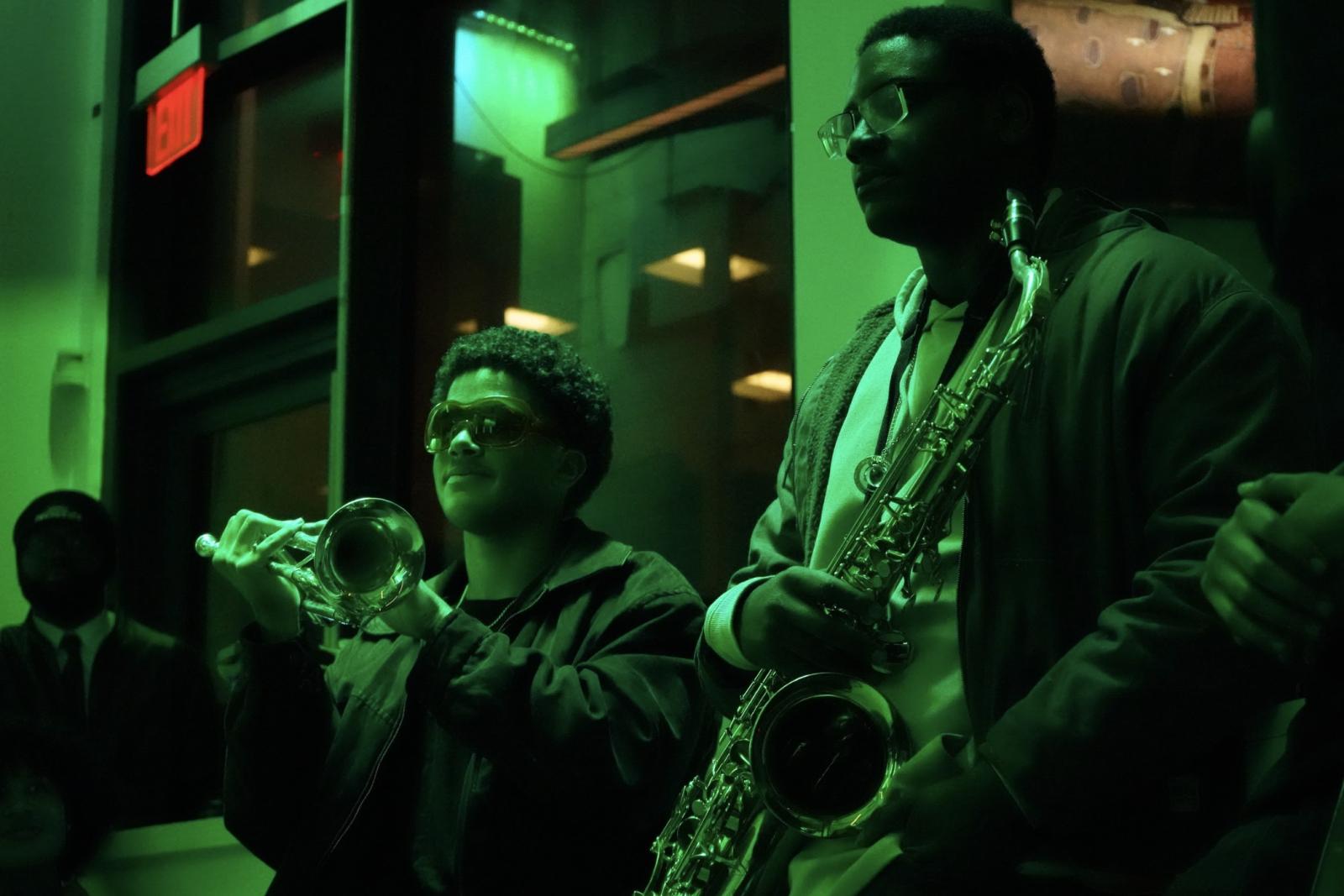 Two male students. the one on the left is holding a trumpet. The person on the right is holding a saxophone. The lighting in the photo has a green hue.