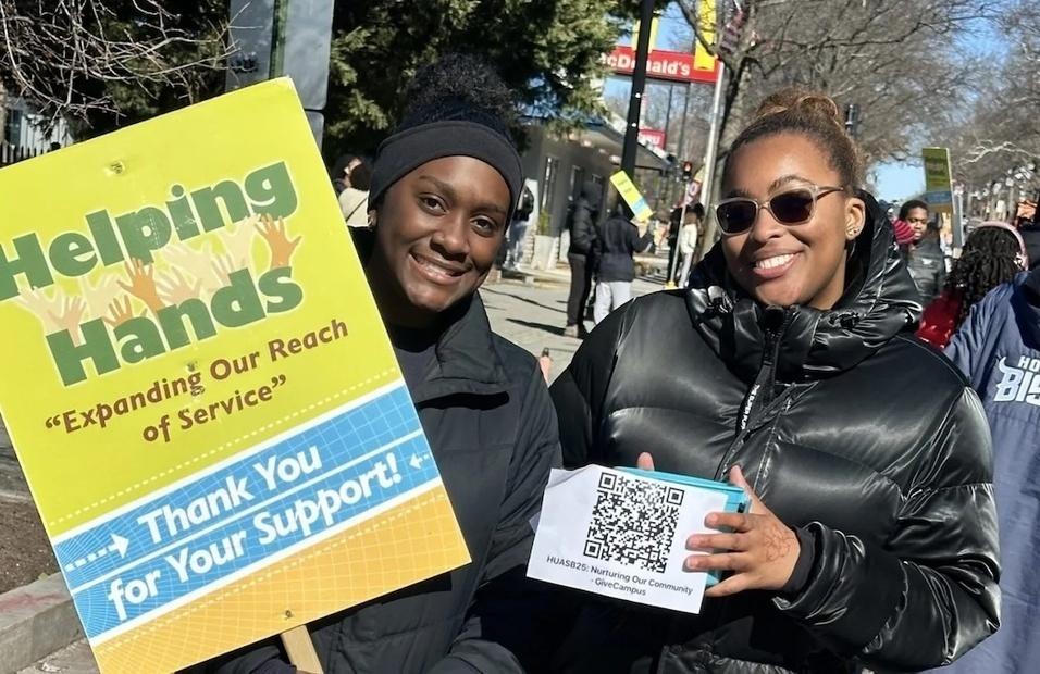 Two howard students outside in winter coats. the student on the left is holding a yellow side that has blue font that says Helping hands. The person on the right is holding a paper that has a QR code on it