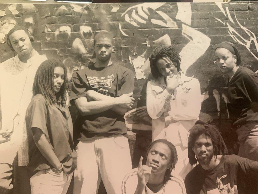 A group of seven people standing up against a wall. The photo is in a sepia tone. 