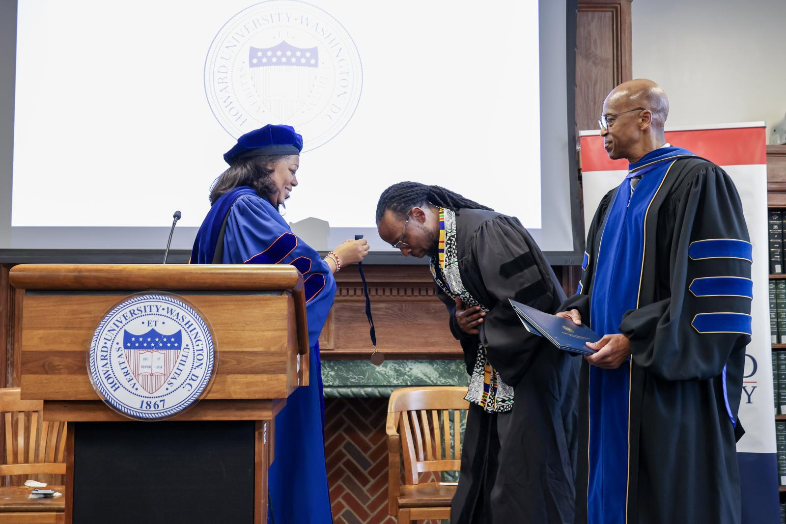 Three people in academic regalia behind a wooden podium. The man in the middle is bowing as the woman on the left is placing a medal over his head. The man on the right is looking to his left at these actions