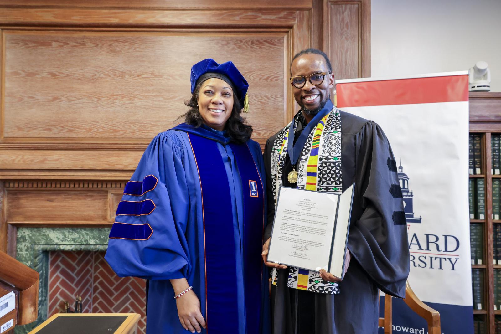 Two people in academic regalia smiling at the camera. The woman on the left is wearing black and blue regalia. The man on the right is holding a piece of paper as he smiles. His regalia is multi colored