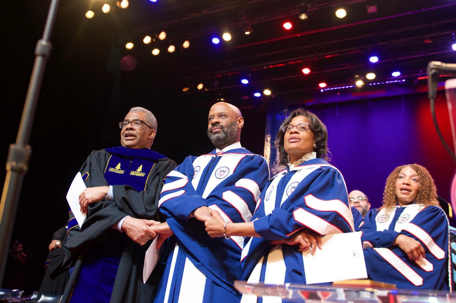Four people holding hands side by side on stage while wearing academic regalia