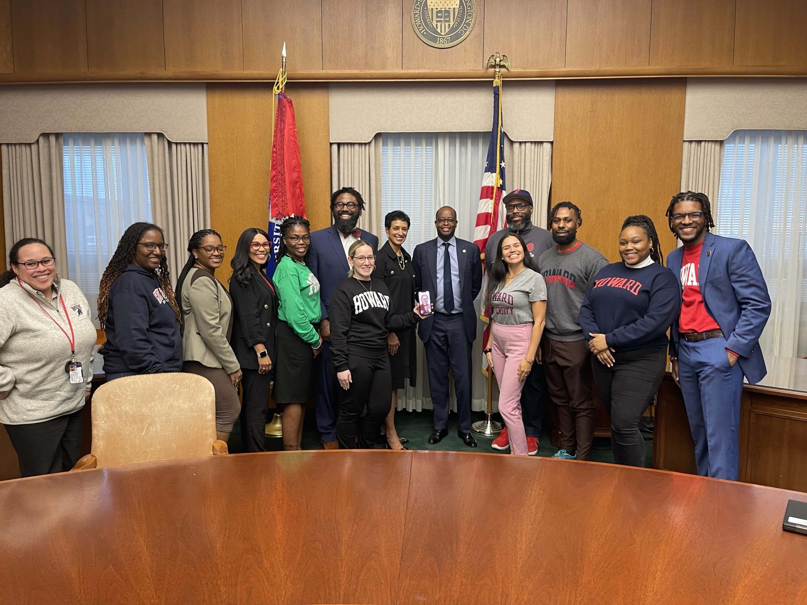 A group 10 people standing side by side in a room. There is a wooden table in front of them