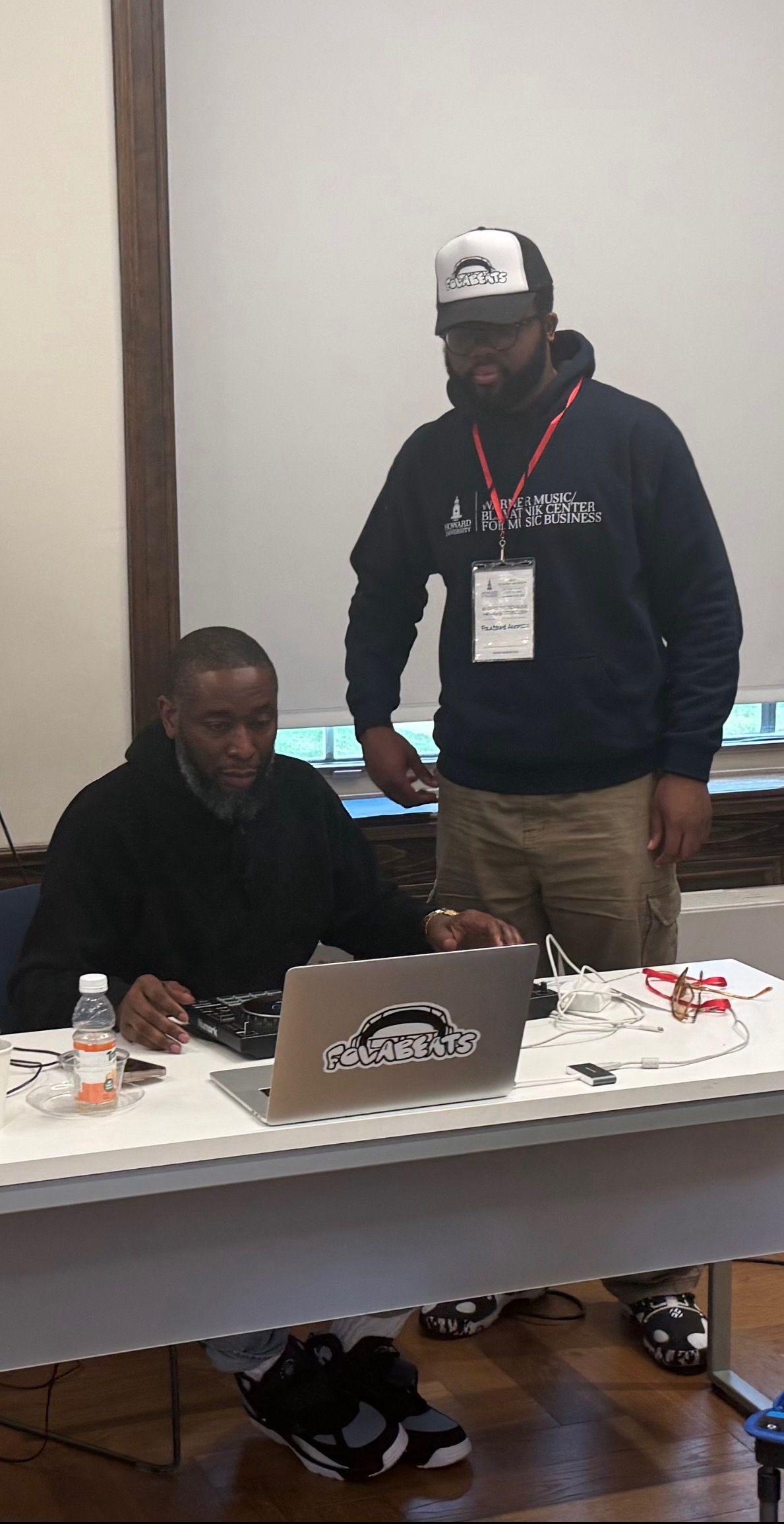 Folajinmi Awofeso on the right and producer 9th wonder on the left. The person on the left is sitting down at a table with a laptop in front of him. The person standing is looking down at person sitting