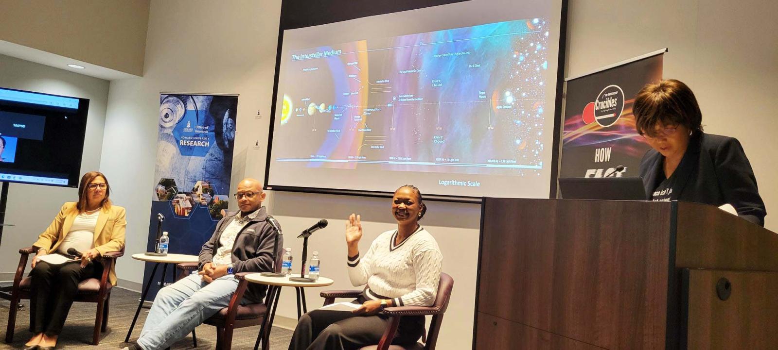 Four people sitting in chairs. There is a person standing behind a podium. There is a projector screen in the background
