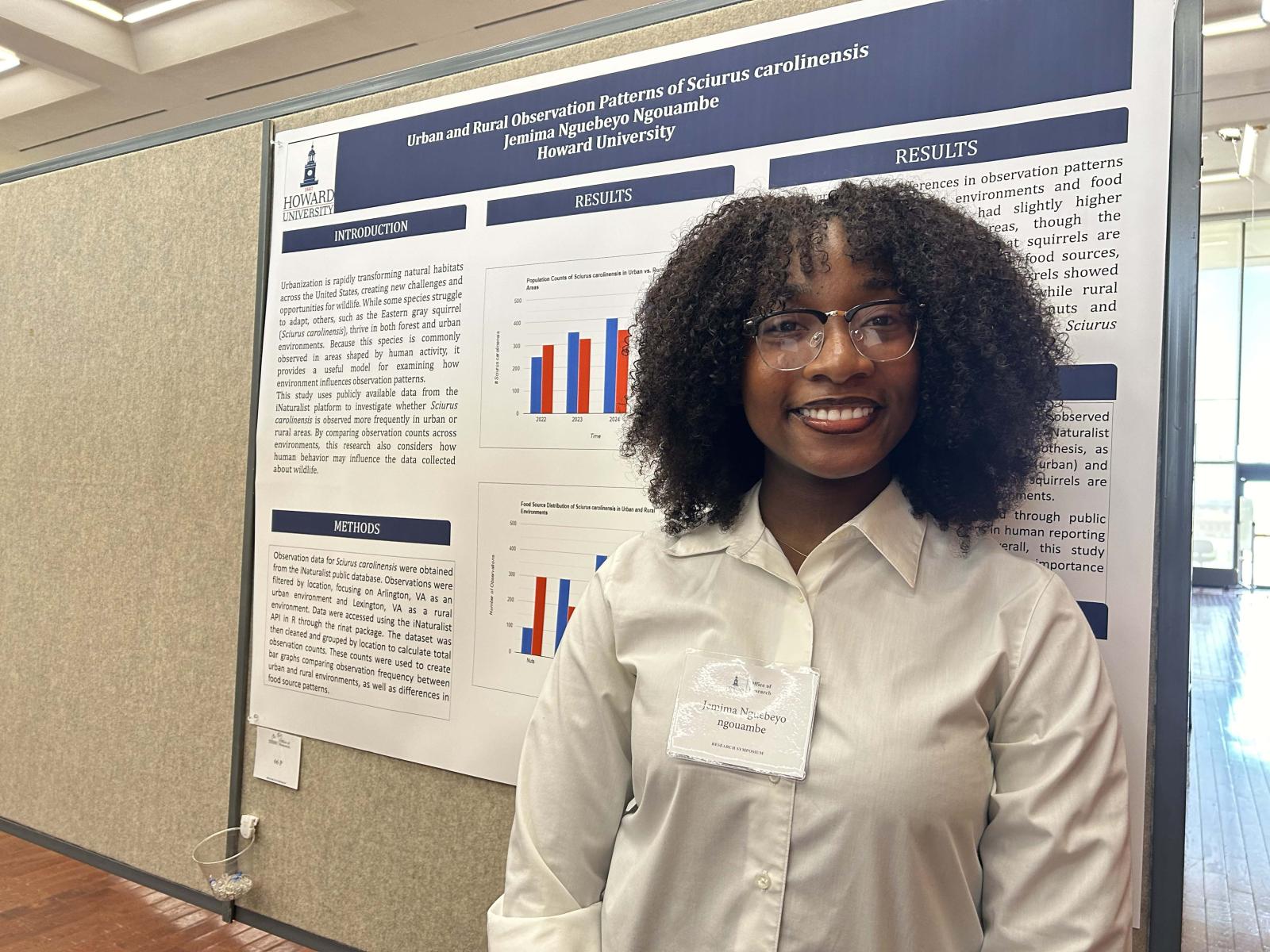 A female college student standing in front of a poster board. she is smiling and wearing a white button down shirt