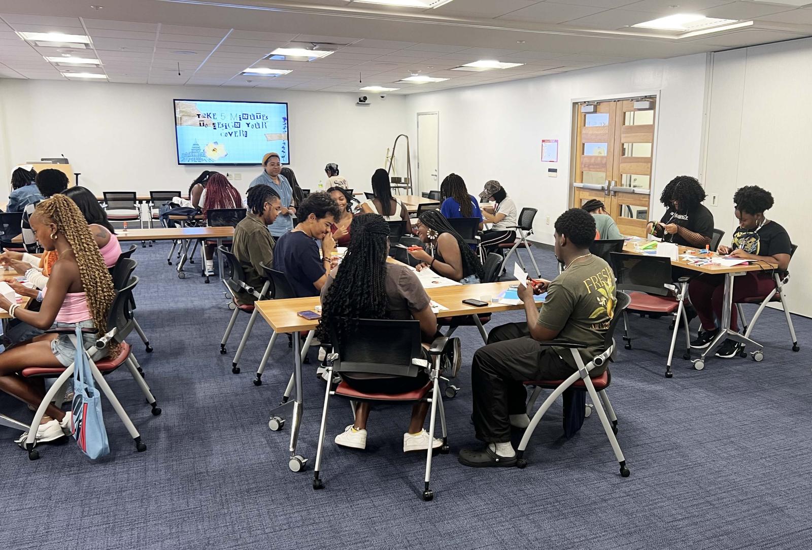 A large group of college students who are all spread out sitting at desks. they are making zines. they have paper, scissors, and other materials in their hands