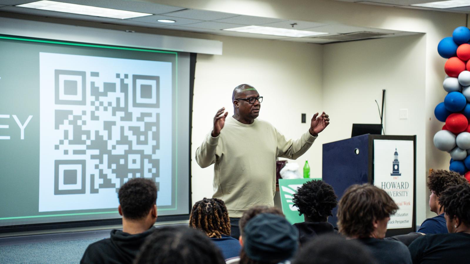 Howard football head coach Ted White standing in front of people gesturing with his hands. There is a large QR code behind him on a projector screen