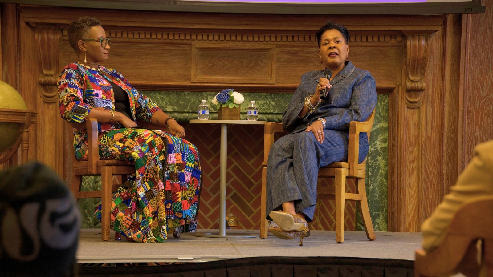 Dr. J. Jarpa Dawuni, wearing colorful African attire, and  Her Excellency Paula-Mae Weekes, wearing a blue outfit, seated on stage at Cramton Auditorium