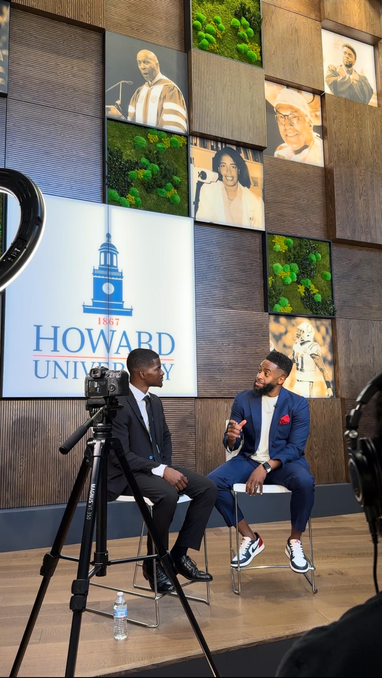Two men sitting on stools in front of a backdrop with many sepia colored photos and the Howard University Logo