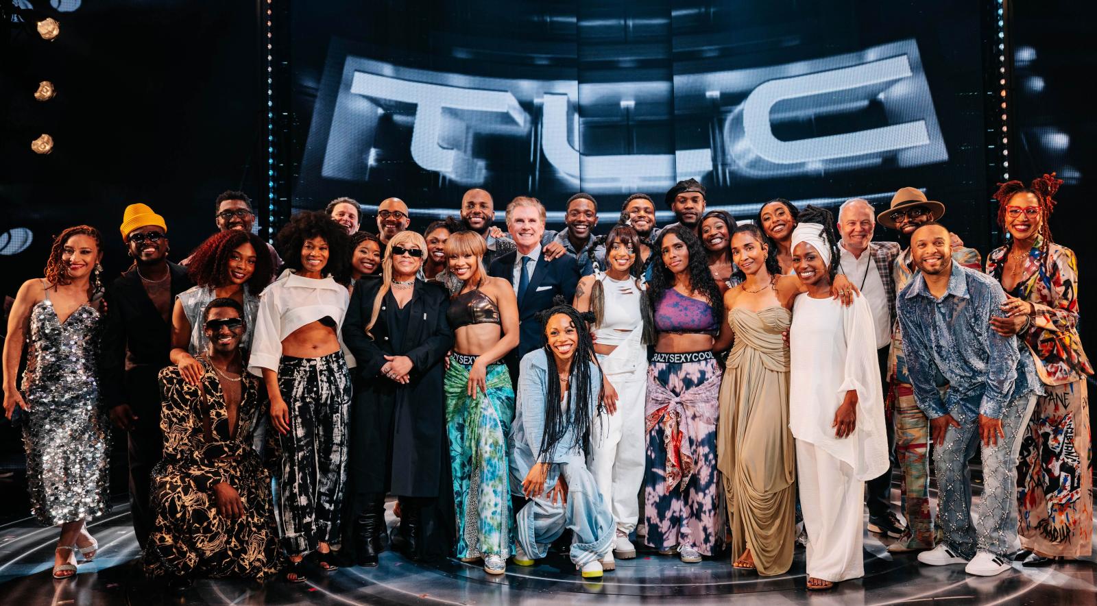 A large group of people standing on a theatre stage for the TLC musical. In the background are the letters TLC