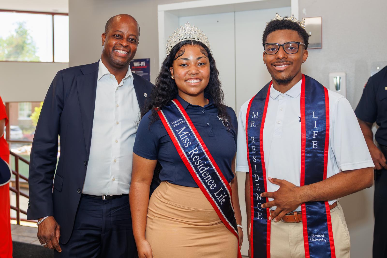 Howard president Wayne Frederick on the left and two howard students smiling. The two students are wearing sashes around their bodies that says Miss Residence Life and Mister Residence Life