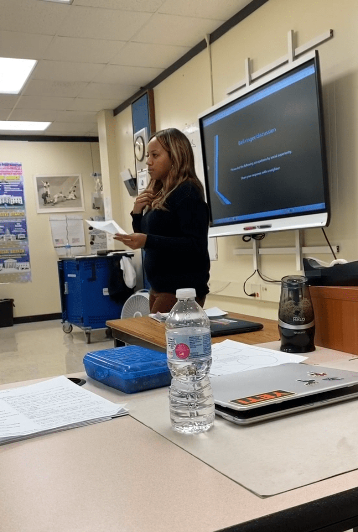 Woman standing in front of a classroom leading instruction. There's a desk in the foreground with items on it such as a water bottle