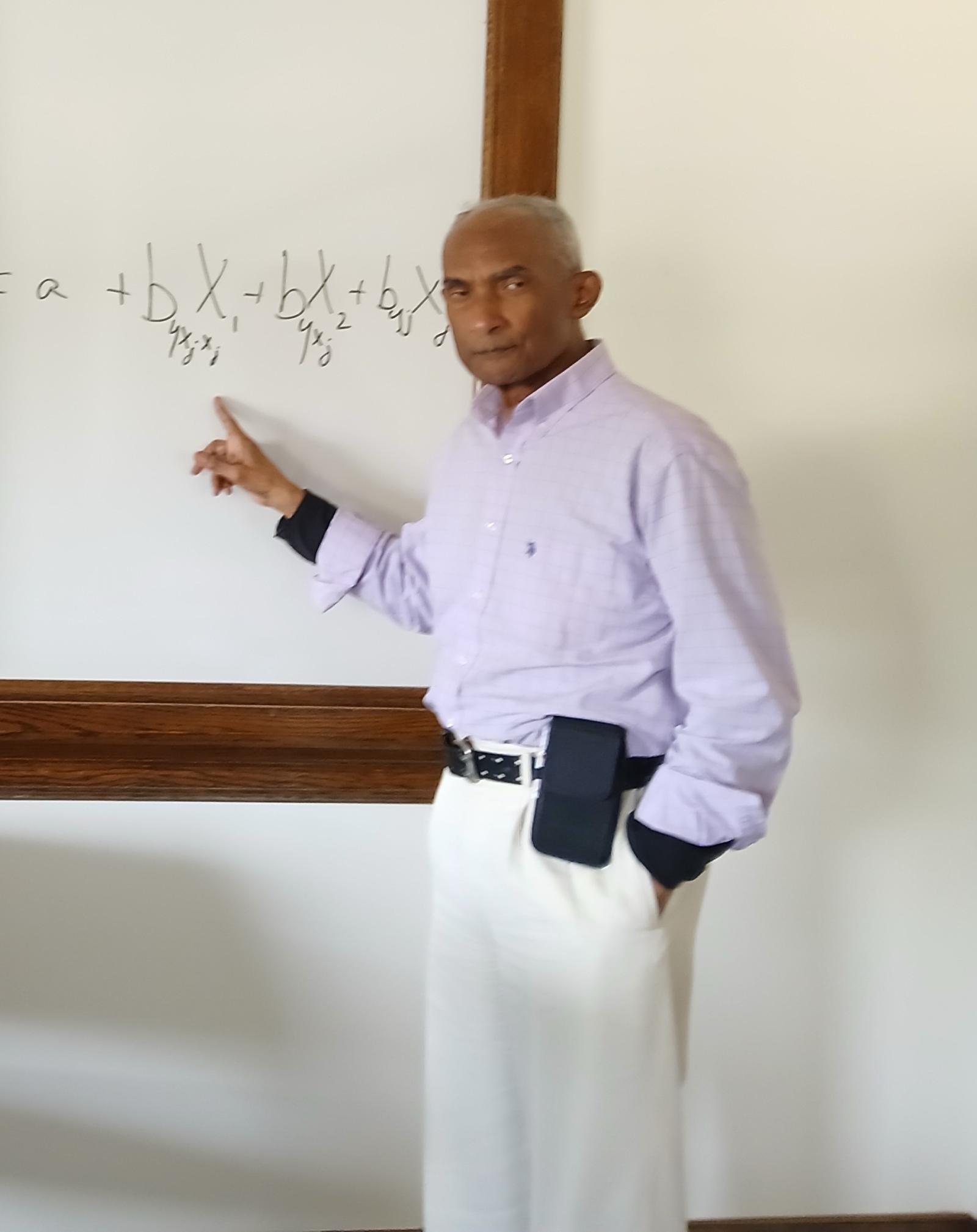 Professor Ron Manuel wearing a purple shirt and white pants standing in front of a chalkboard