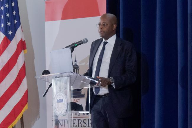 Dr. Wayne A. I. Frederick wearing a dark suit speaking at a podium next to the American flag delivering the State of the University address in 2025